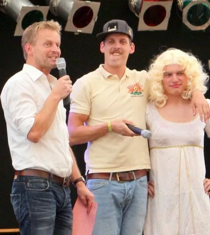 Moderator interviewt zwei Teilnehmer, einer davon mit blonder Perücke und Kleid